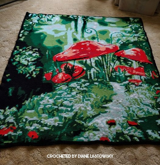 A square crocheted tapestry or rug featuring a whimsical forest scene with large red mushrooms, green foliage, and a light gray path, laid flat on a beige carpet. Text at the bottom reads 'CROCHETED BY DIANE LASTOWSKY'. Fairytale Mushroom. Design by Geeky Graphghans