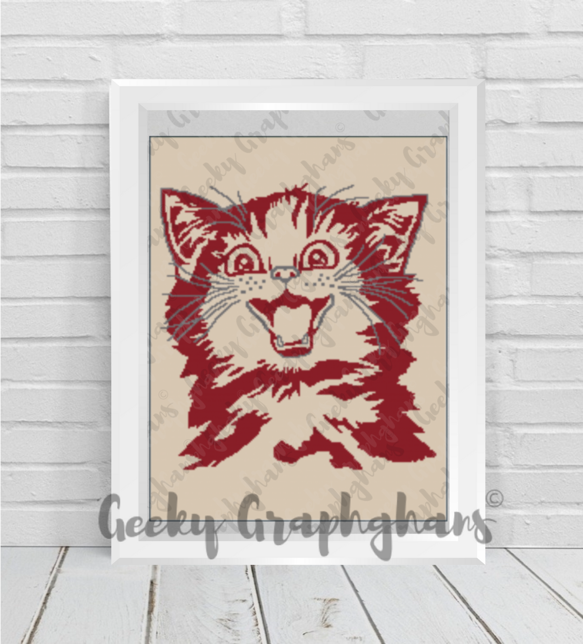 A crochet graphghan pattern depicting a stylized happy kitty face in red and orange on a beige background, displayed in a frame.