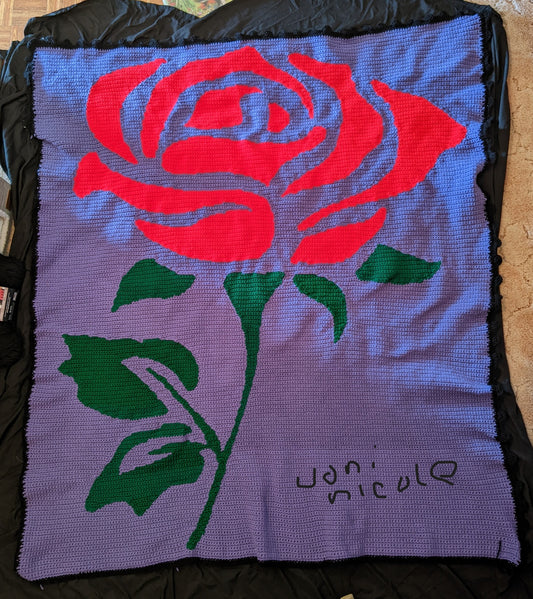 Crocheted blanket with a red rose design on a purple background, signed 'Joni Nicol'.