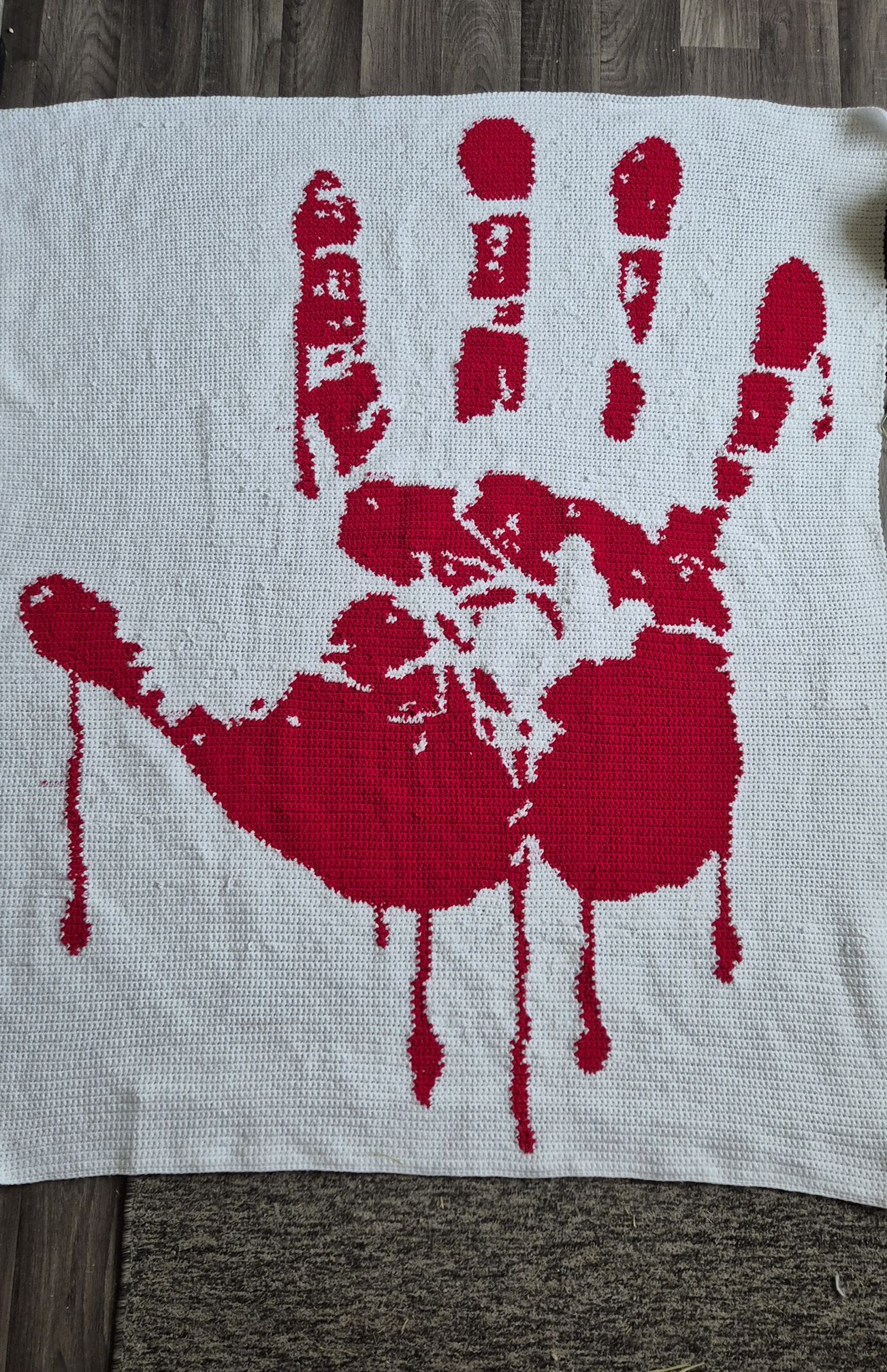 White fabric with a red handprint and blood-like design on a wooden surface. Bloody Hand crochet graphghan crocheted by Catherine Worrall. Design by Geeky Graphghans