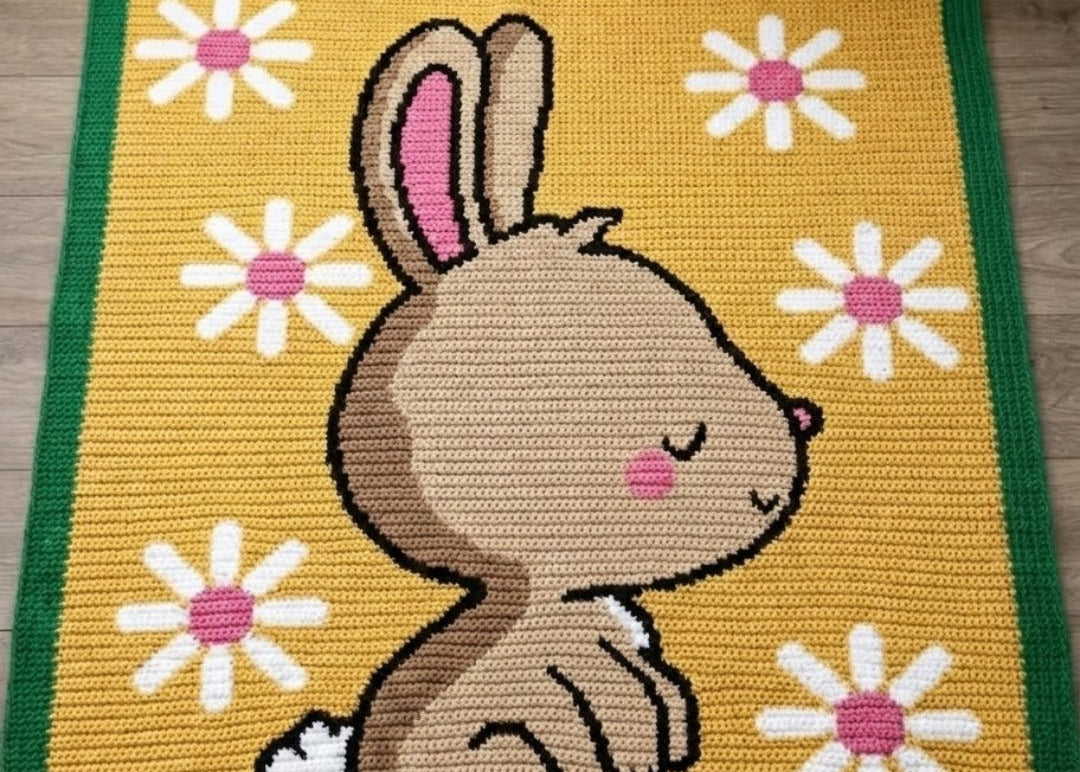 Crocheted blanket with a bunny design and flowers on a yellow background, featuring 'Geeky Graphhans' branding.