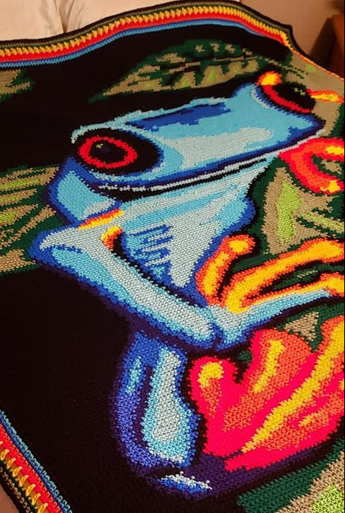 Colorful crocheted blanket with a frog design draped over a wooden surface.