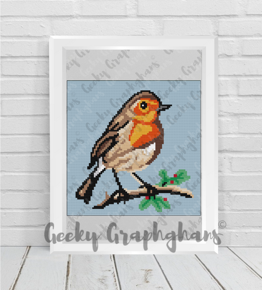 Cross-stitched robin on a branch with holly leaves in a white frame, on a light wooden floor. Crochet Graphghan pattern by Geeky Graphghans