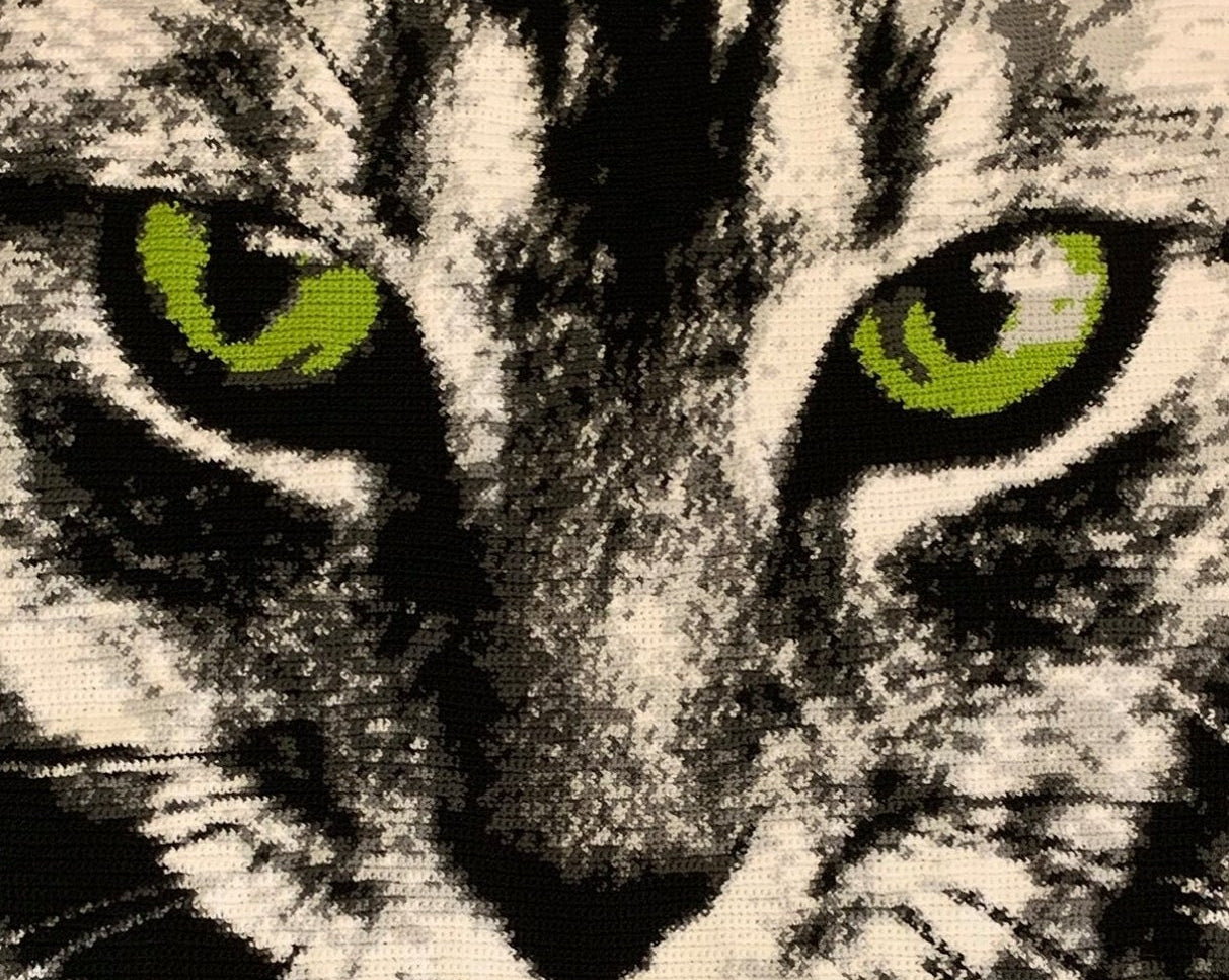 A close-up woven or embroidered cat portrait with bright green eyes, black and white fur, and a pink tongue sticking out, shown against a neutral textured background. Text at the bottom reads 'Daniela Navarro von Starck 1,05m x 1,40m'.